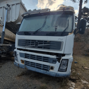Cabine Volvo FM branca vista de frente e baixo, com o basculante de uma caçamba ao fundo. Mostra danos frontais.