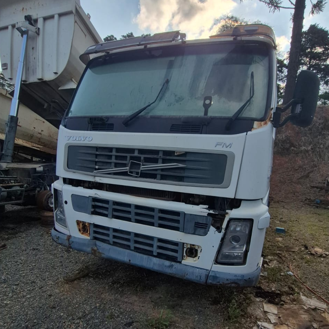 Cabine Volvo FM branca vista de frente e baixo, com o basculante de uma caçamba ao fundo. Mostra danos frontais.